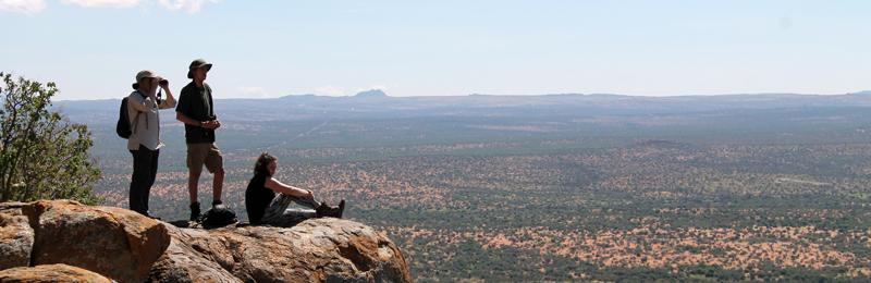 This image shows three students on the african field course. They are standing on top of a large rock looking into the distance. Two of the students are standing while the other is sitting.