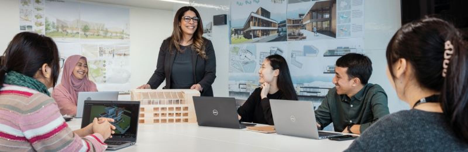 A group of students sat at a desk with their laptops, with a academic in the middle showing them a model of a building. On the walls are architectural designs.