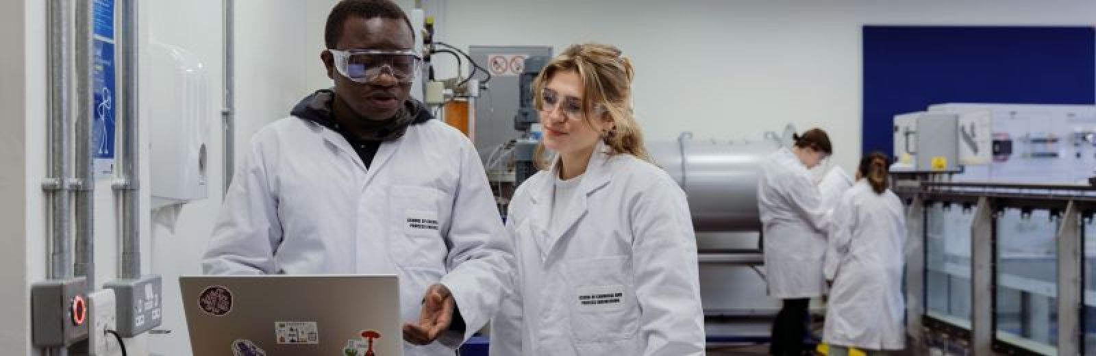 Students in a chemical engineering, renewable energy lab. Two students are in the foreground stood at a table with a laptop, dressed in white labs coats and safety glasses.