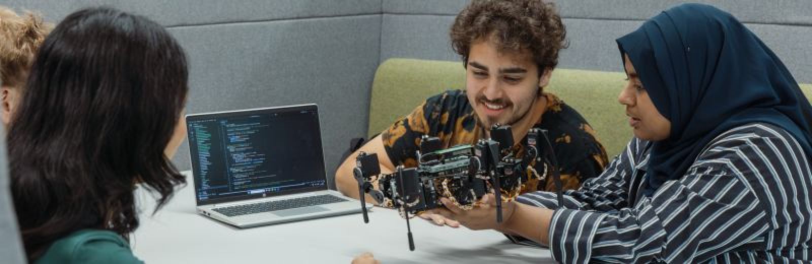 Three students sat in a booth, with a eight legged robot and a laptop displaying code that runs the robot. Two students are handling it carefully.