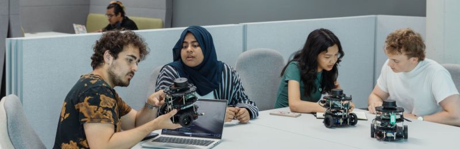 A group of four students sat a desk with small robots controlled by AI, via the laptop on the desk.