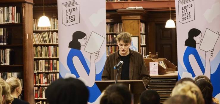 A student stands at a lecturn in a traditional looking library setting. He speaks into a microphone to the crowd of people sitting in the foreground. Behind him are two banners that read Leeds Lit Fest 2023.