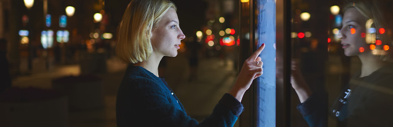 Young lady with short blonde hair standing in the street at night looking at and interacting with an artificial intelligence billboard with her reflection on the screen.