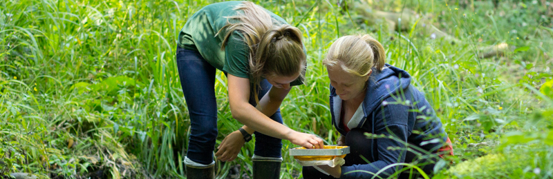 This picture shows to female students studying water collected from a river. One of the girls is crouching while the other stands next to her.