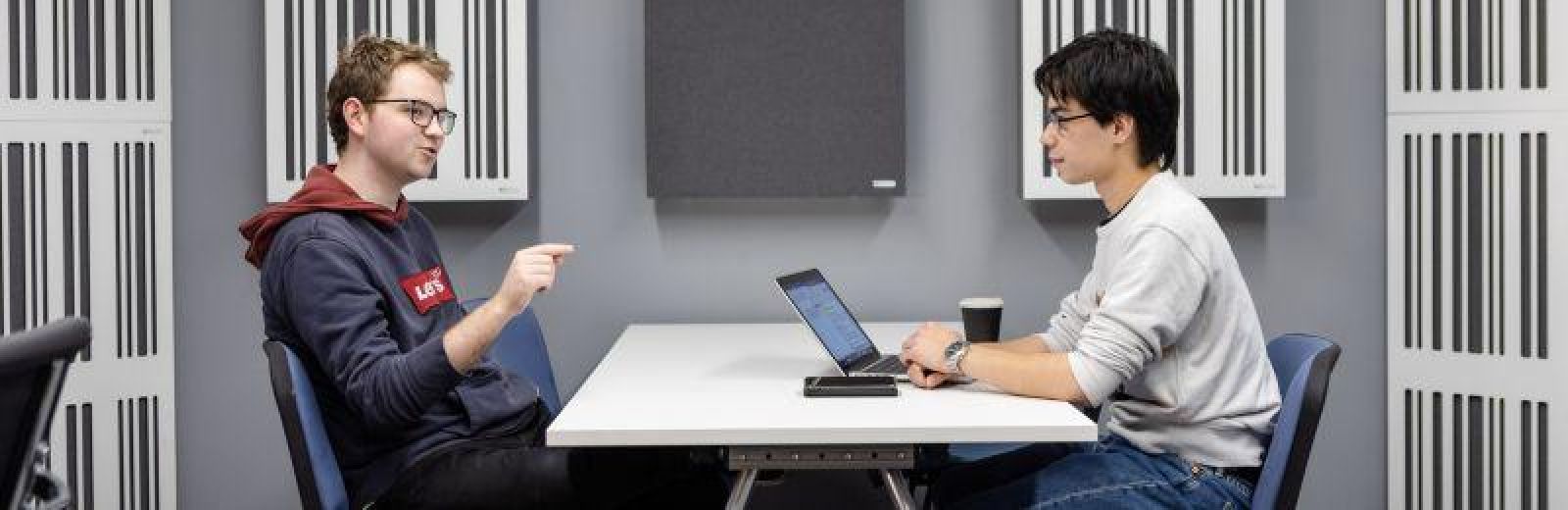 Two Music students in the Music Psychology Lab. They are sat at a table facing each other discussing a research project.