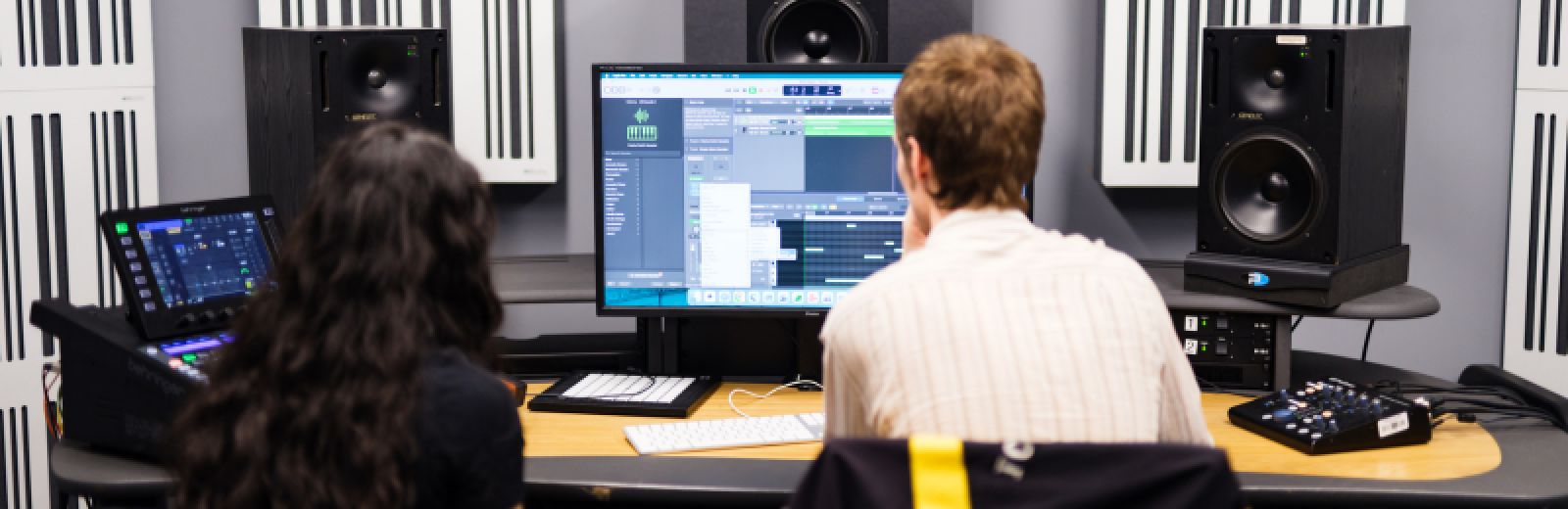 Two students sit at a desktop computer, with a large monitor displaying music software. Three large black speakers surround them behind the screen.