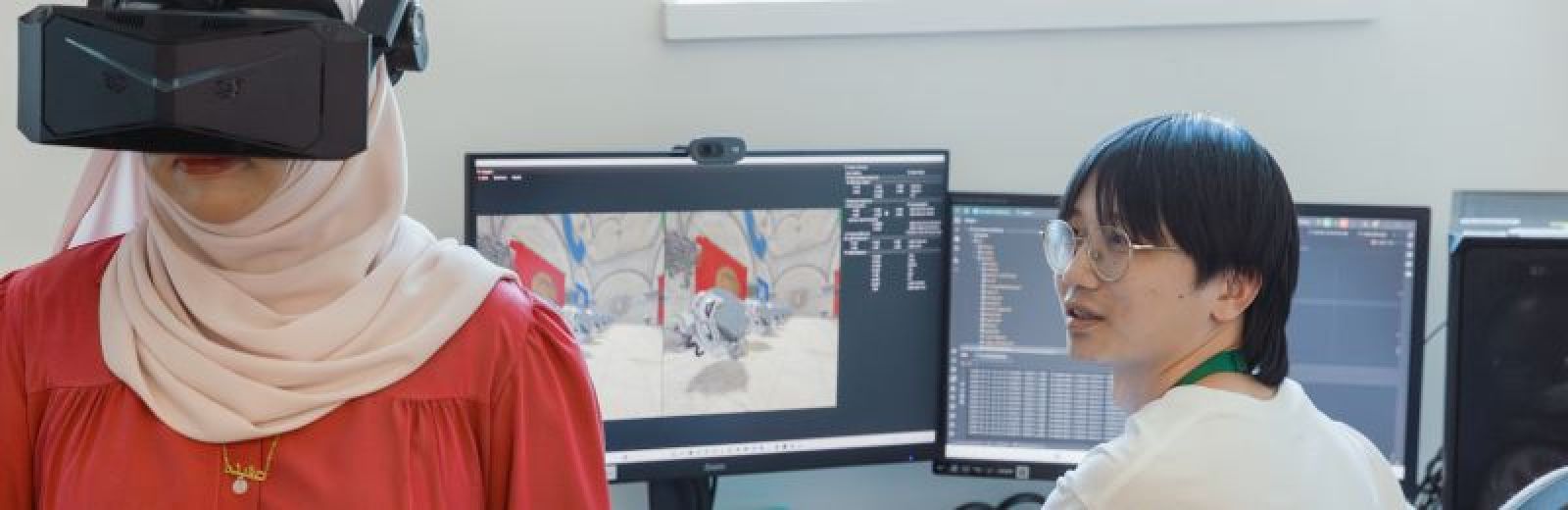 Two students are in a games and VR lab. One student is sat down at a desktop computer, a virtual environment displayed on the monitor. The other student is stood next to the desk, wearing a black VR headset, obscuring their face.