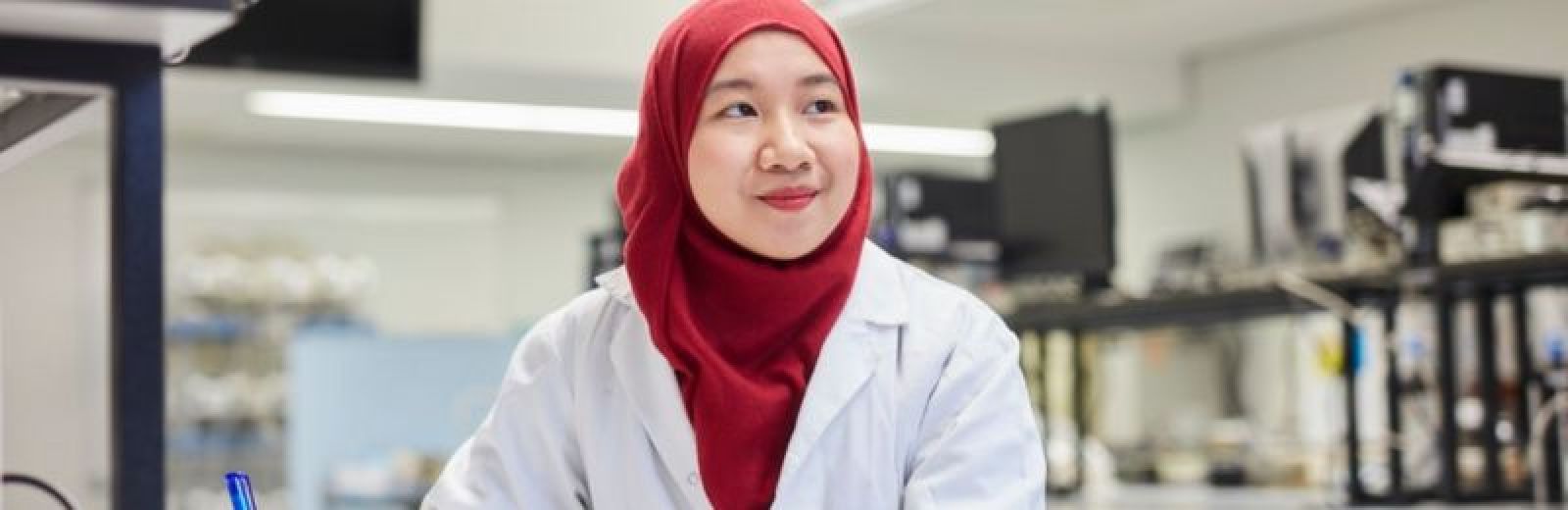 A student sat at a work bench in a biomedical lab, wearing a white lab coat and red headscarf.