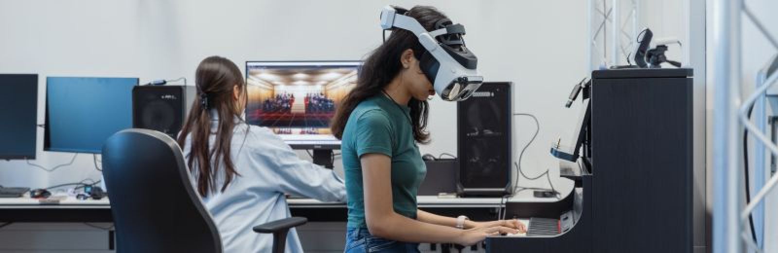 Two students in a computer virtual reality (VR) lab. One student has a VR headset on and is sat down at a piano. The other student is sat behind them at a computer, the visual of a concert hall is displayed on screen.