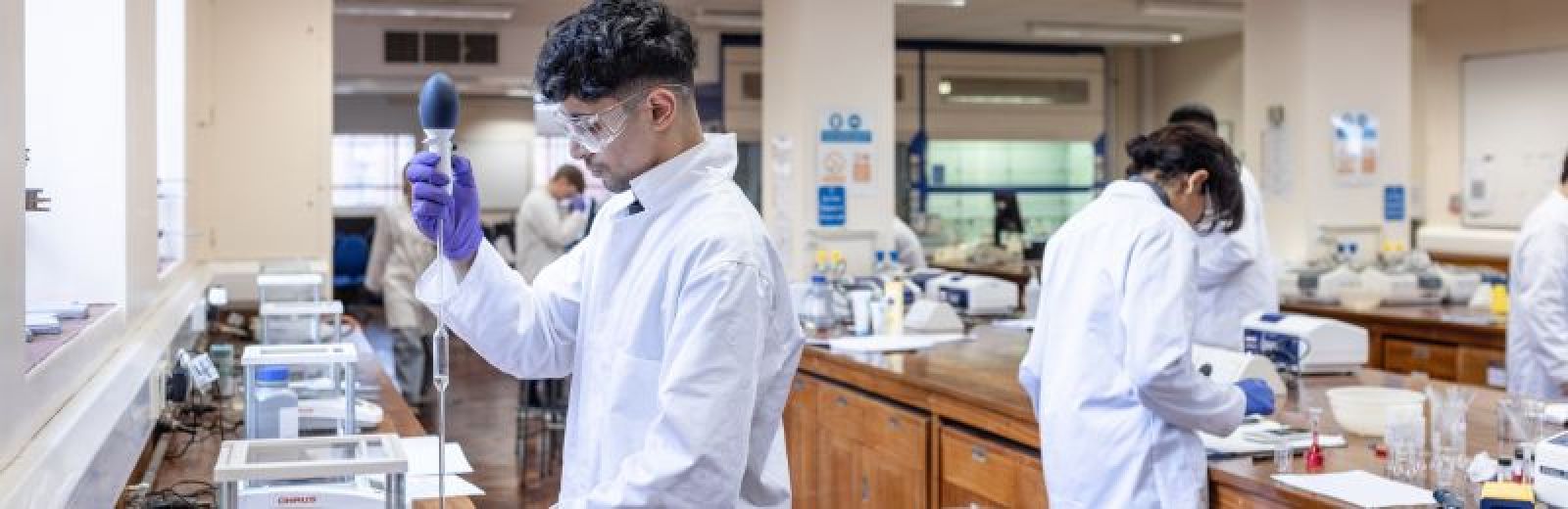 Students working in a chemistry lab at the University of Leeds. The students are dressed in white lab coats, and protective eye wear.