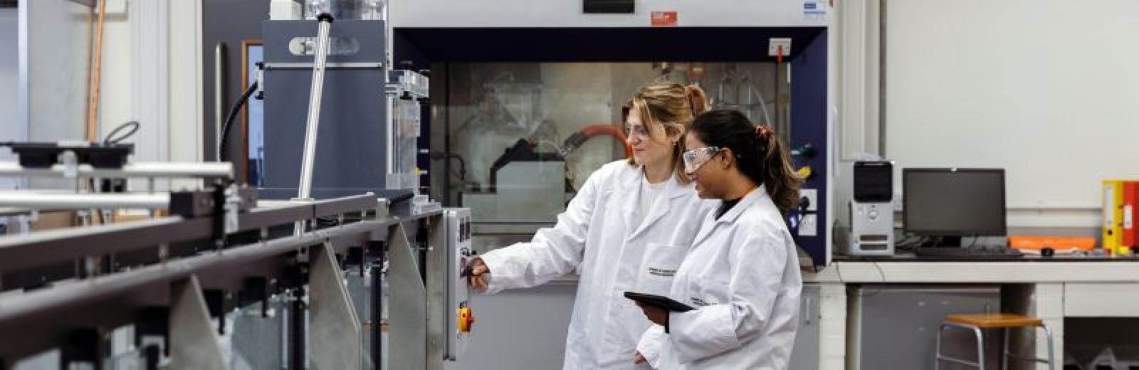 Two students stood in a renewable engineering lab, wearing white coats. They are looking at the controls on a wave machine, behind them is a fume cupboard.
