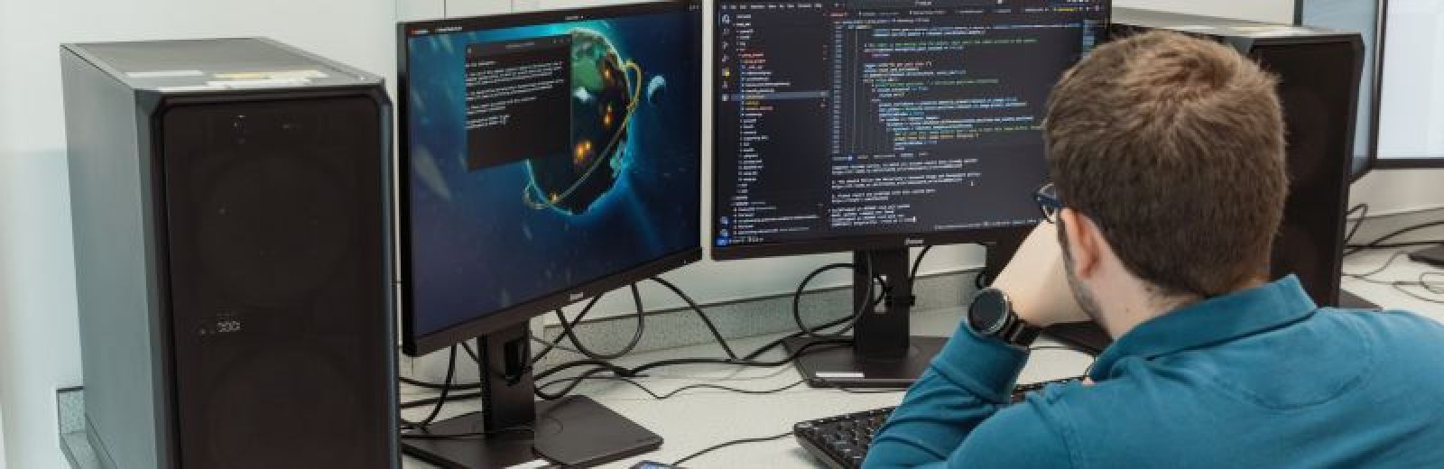 A computer science student is sat at a desktop computer, that has two screens. Both screen shows windows of coding.