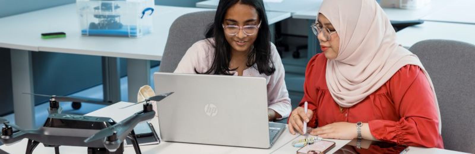 Two students sat working at table with an open laptop and a drone in front of them.