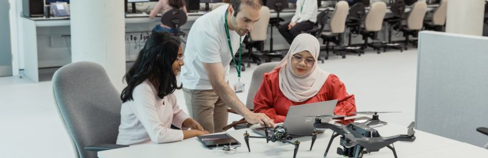 A photograph of two students and a academic in a computer science lab. The students are sat a desk with two different sized drones and a laptop. The academic is showing the students something on the screen. Behind them is a row of desk and computer chairs