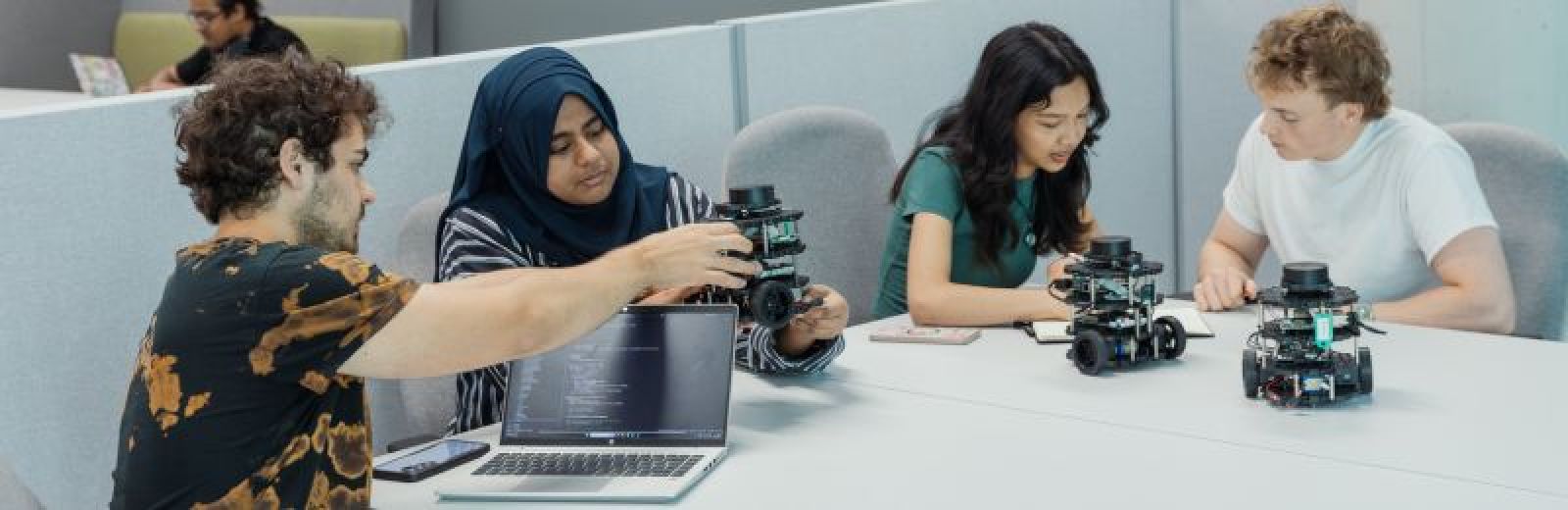 Four students sat a table working on three AI powered robots. One student has an open laptop in front of them displaying programming code that runs the robot.