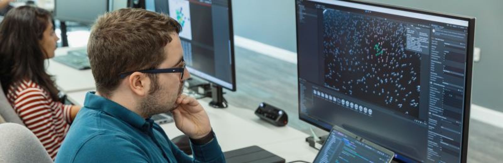 A photograph showing two students each sat at a desk looking at a computer monitor. each screen displays a different computer science project.