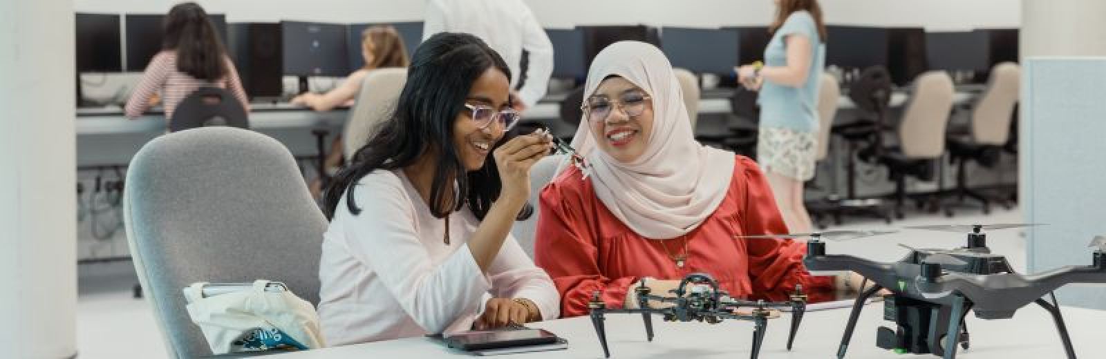 Two computer science students sat at a desk in a computer science lab. The students are looking a three different sized drones in front of them on the table. Behind them are other students sat working at desktop computers.