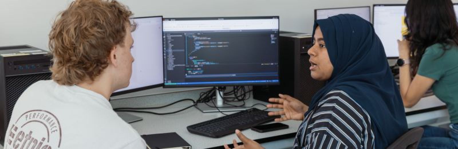 Two students sat a desktop computer with two monitors showing programming code.