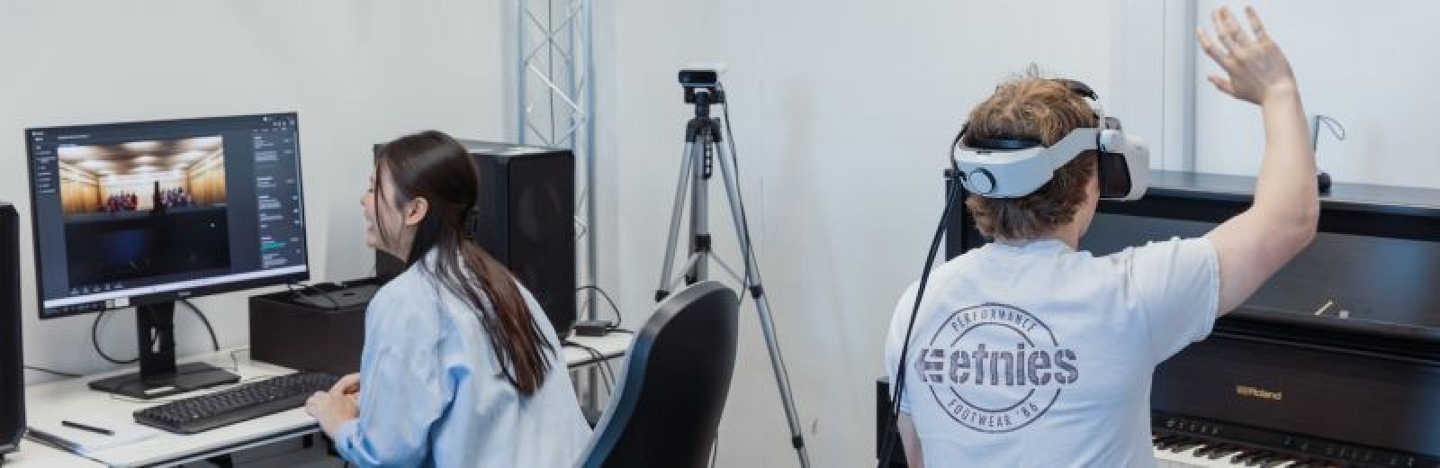 Two students in a High-Performance Graphics and VR lab. One student is sat at a desktop computer, the screen displaying a VR auditorium. The second student is sat at a piano wearing a VR headset, right arm in the air waving to a virtual audience.