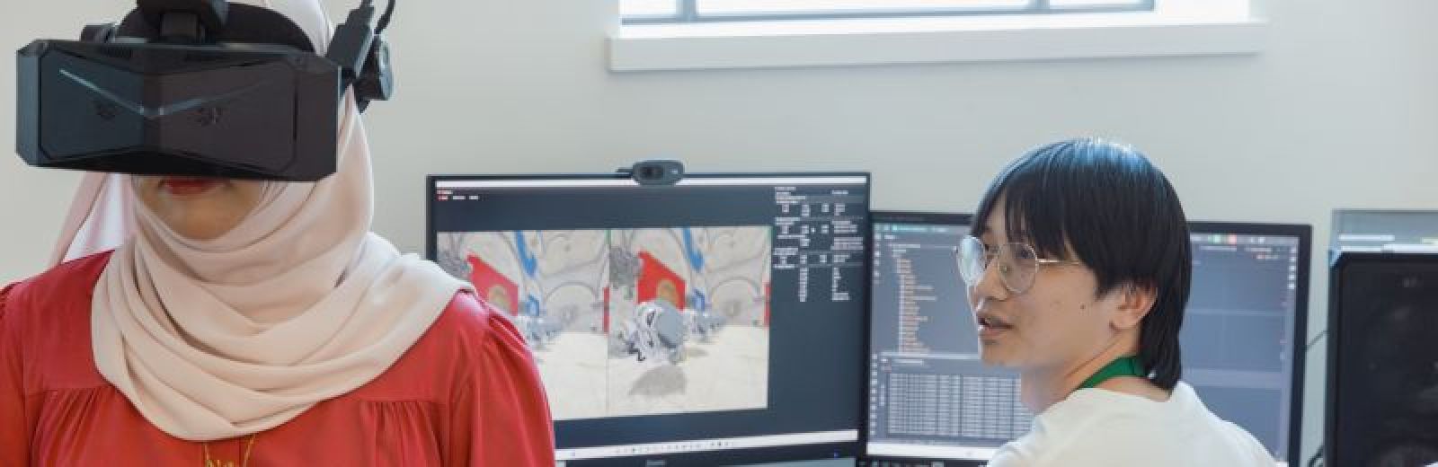 Two students in a VR lab. One is sat at a desktop computer, the screen displaying a colourful virtual environment. The other student is stood wearing a VR headset.