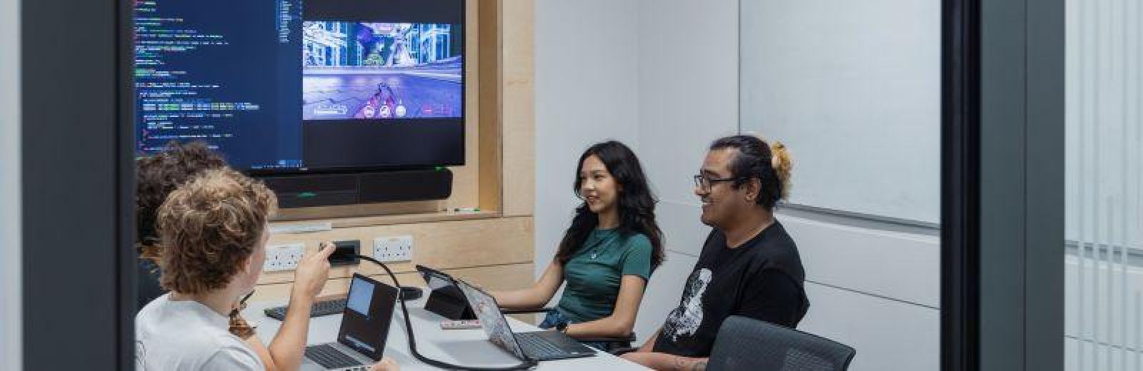 A group of students sat around a desk in small meeting room. There is a big screen displaying a split of code and the rendered game from the code.