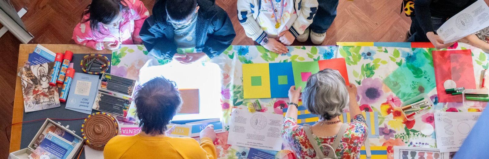 People of different ages gathered around a table interacting with coloured paper, pencils and other craft materials.