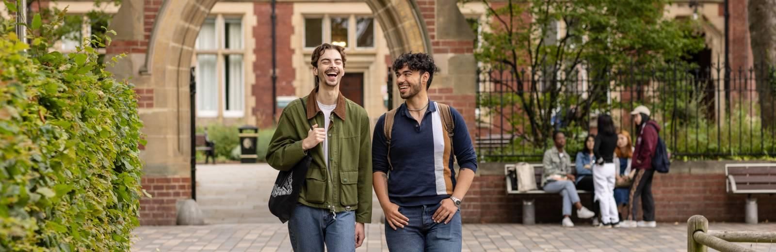 Two students walk through campus side by side, they are laughing. In the background another group of students sit at a bench, and the redbrick arch of Clothworker's Court can be seen.