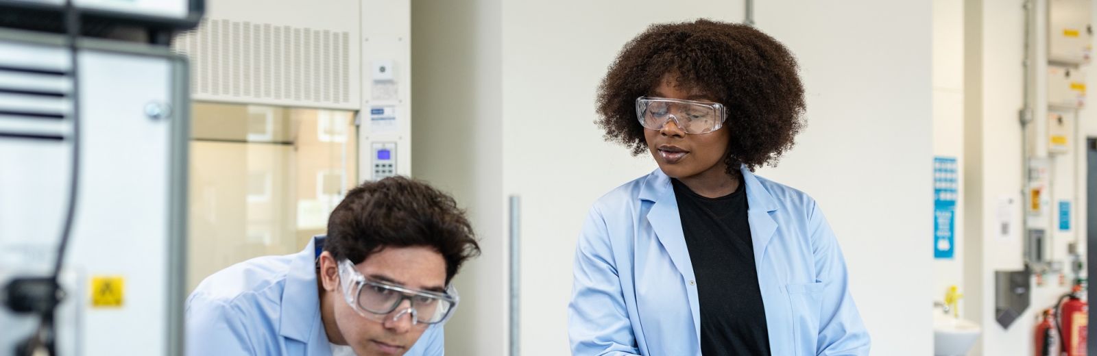 Two students wearing blue lab coats and clear safety glasses, stood in a chemical engineering lab.