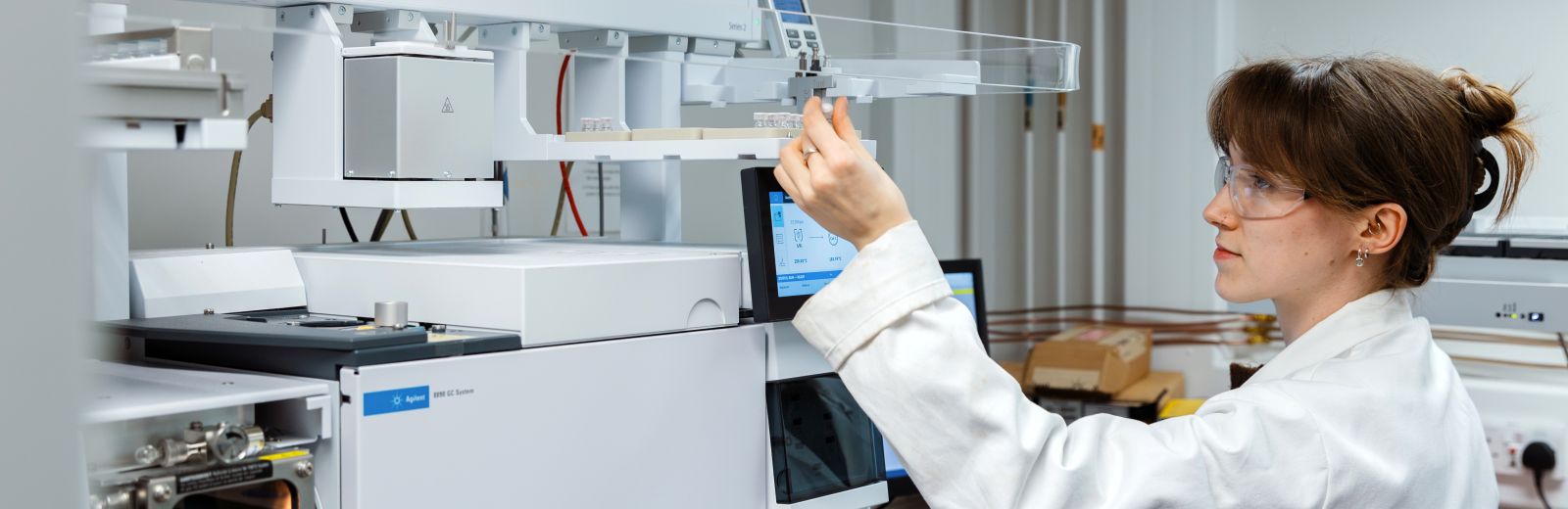 A student, wearing a white lab coat, is loading a sample in to a piece of equipment in a digital chemistry lab.