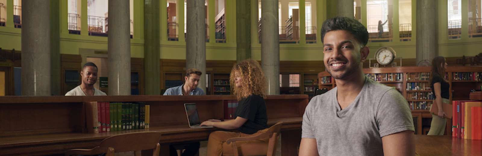 A male student sits in the Brotherton Library and smiles towards the camera. In the background other students can be seen working in the library.